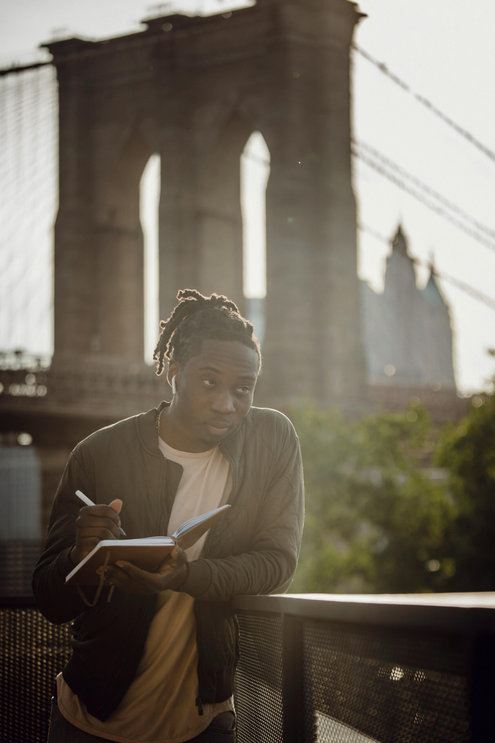 pexels-photo-4559961-4559961 A young man writes in a notebook on a sunny day with the Brooklyn Bridge in the background.